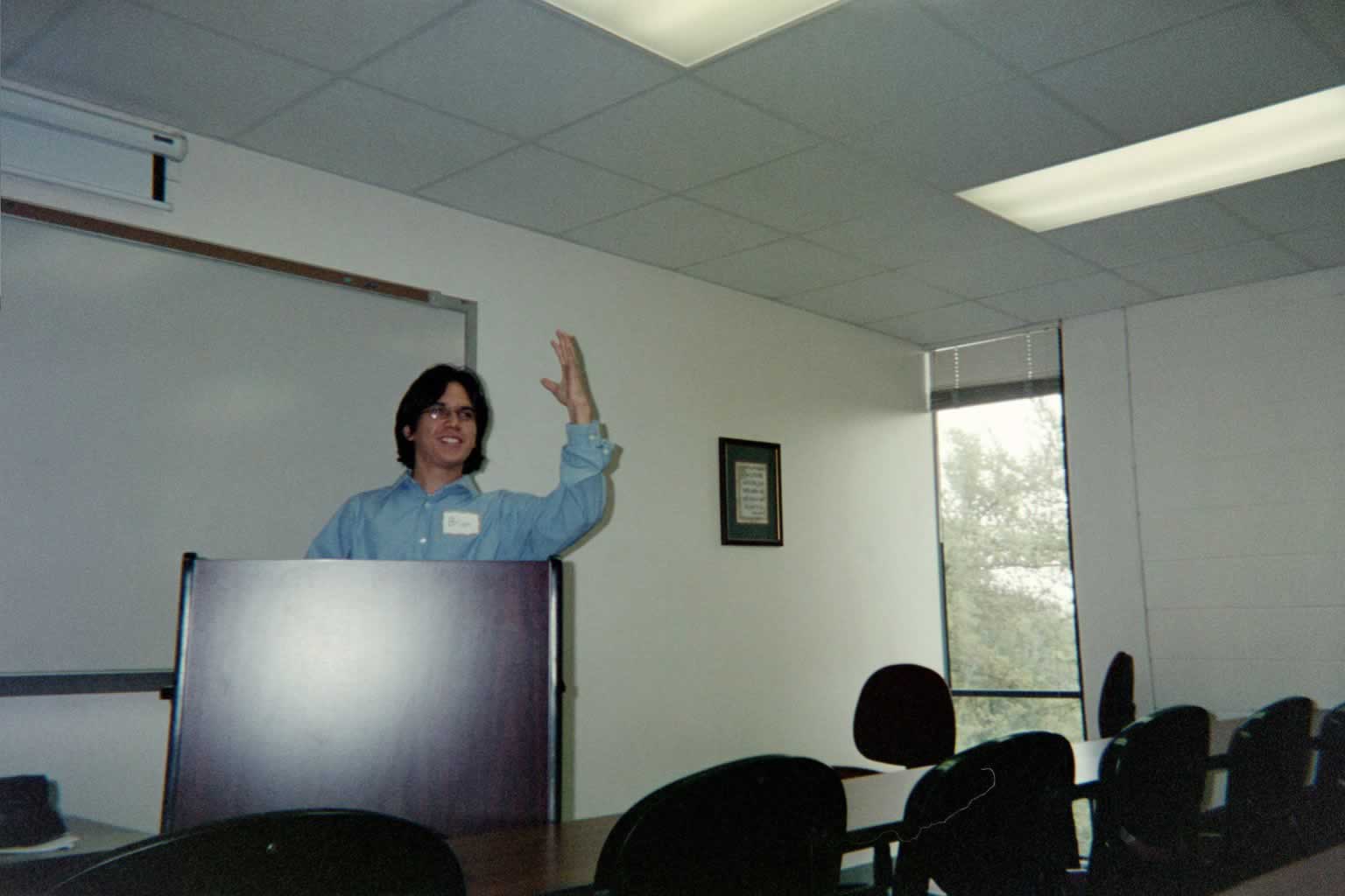 picture of a man in glasses standing behind a podium smiling with his hand held up