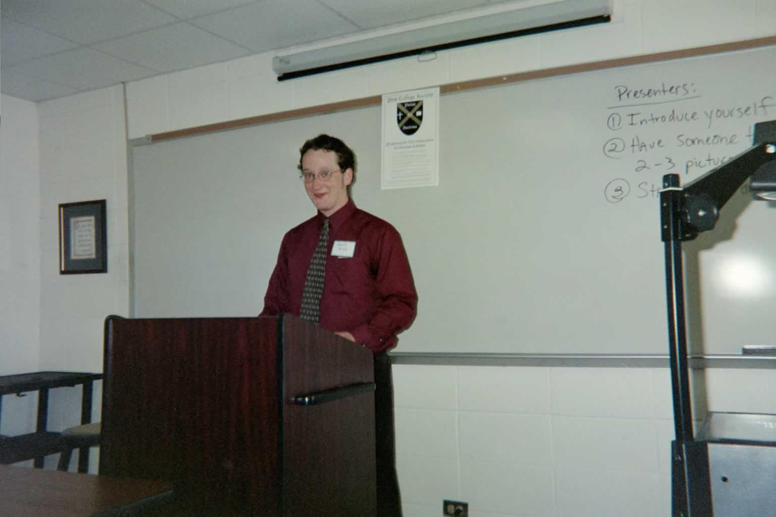 picture of a man wearing glasses and a red shirt standing behind the podium smiling