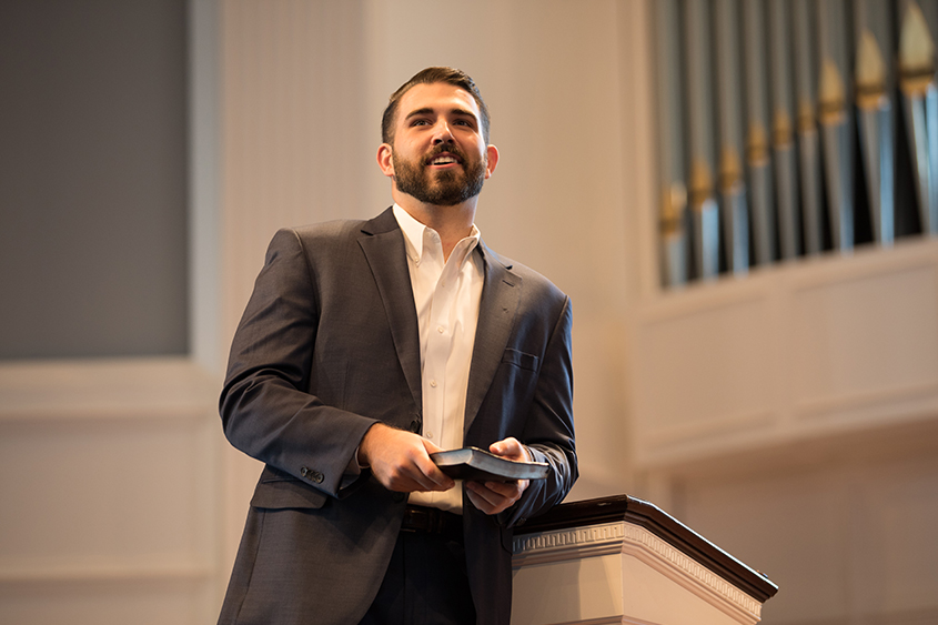 A man stands in front of the podium in the chapel holding a Bible