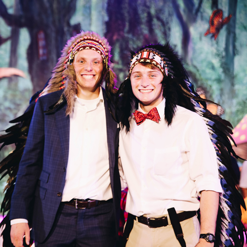 Two guys during Mr. Big Chief posing with headdress on and one is wearing a red bowtie