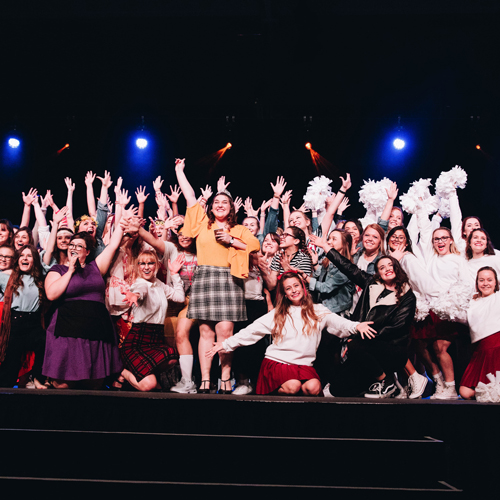 A large group of girls posing on stage with their hands raised in the air at a DBU student event