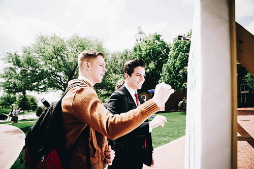 Two guys outside looking at a sign, the one on the left is wearing a brown coat and the one on the right is wearing a tux