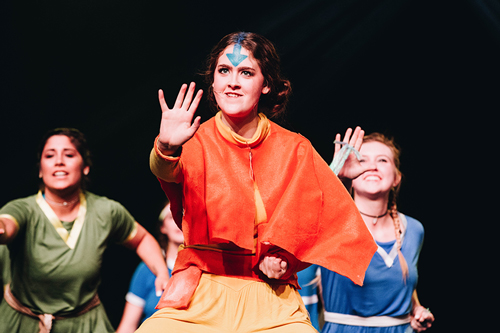 A girl wearing orange holding her hand up, with a blue arrow painted on her forehead.