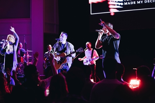 A band singing, the photo is darker and there are purple lights in the background