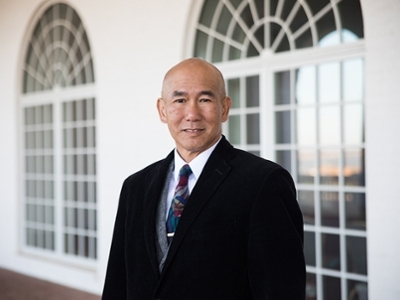 Russel Chun is standing in front of a set of windows smiling.