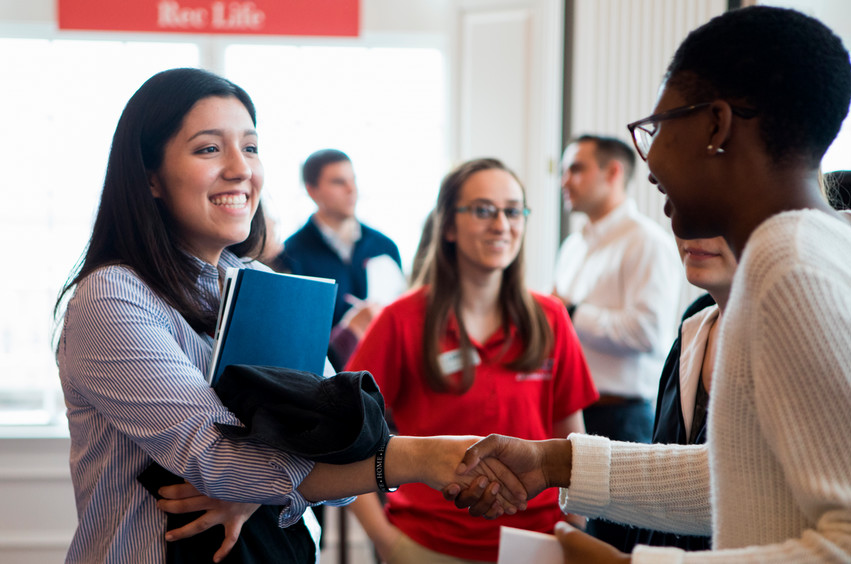 Two students smiling and shaking hands with a group of people in the background